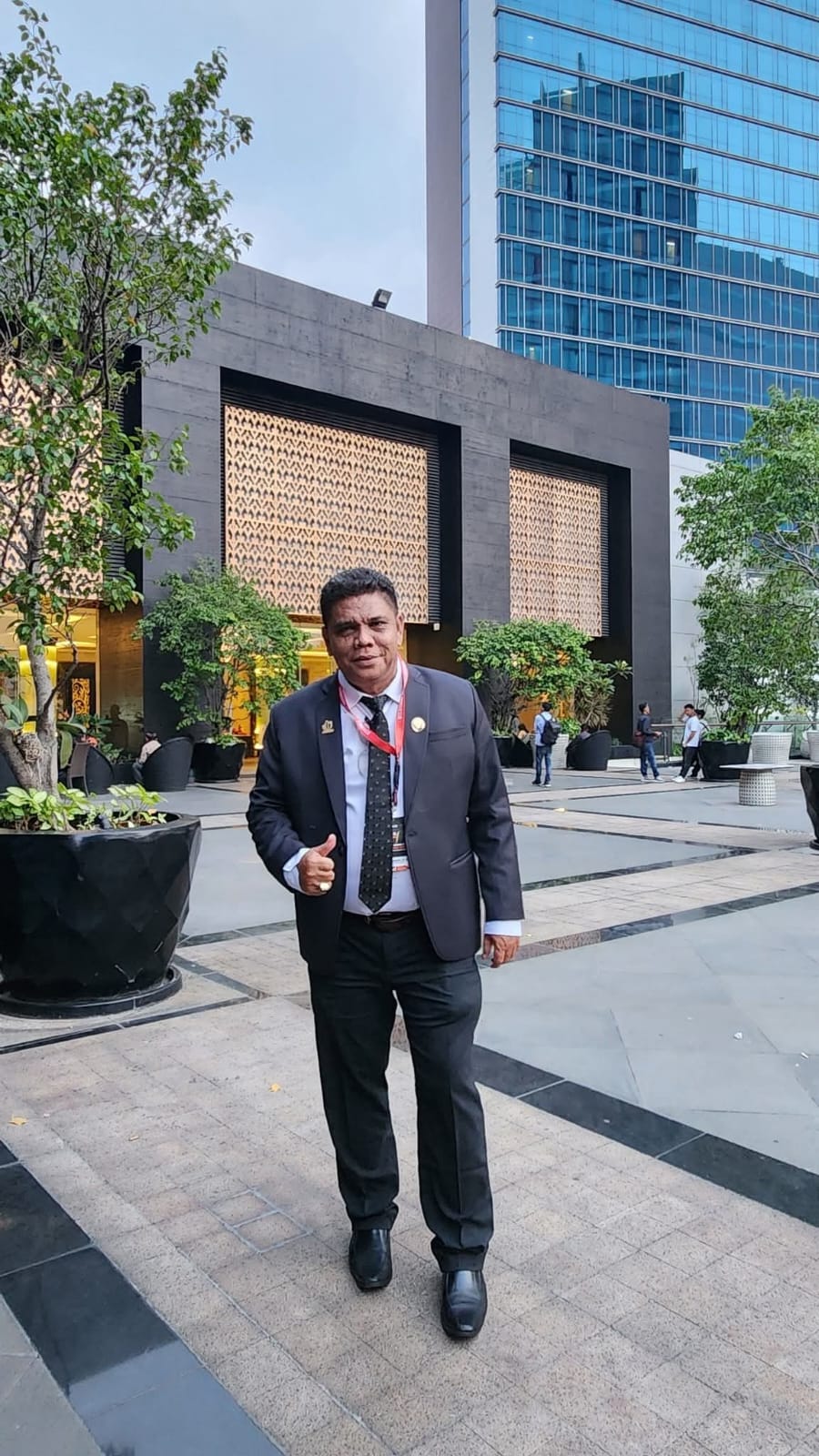Bapak Raymond Simamora, S.H., M.H. berpose mengenakan setelan jas formal dengan latar belakang area gedung perkantoran modern.