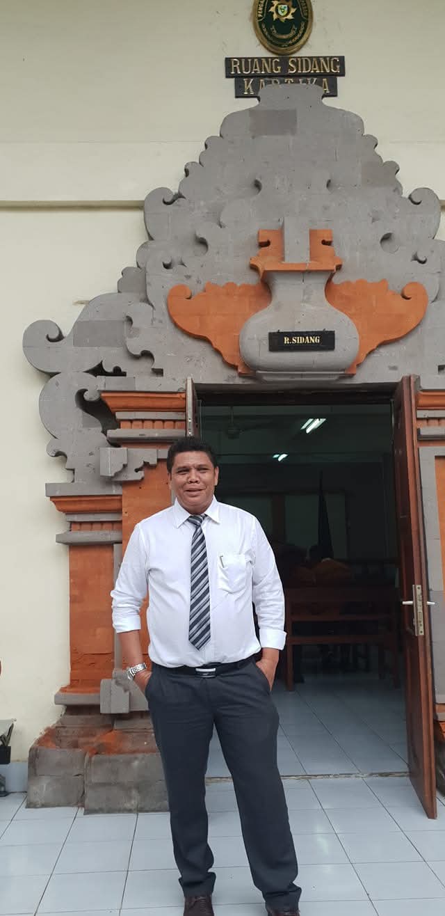 Potret Raymond Simamora, S.H., M.H. saat berada di depan Ruang Sidang Kartika di sebuah instansi pengadilan, dengan latar belakang arsitektur bergaya Bali yang khas.