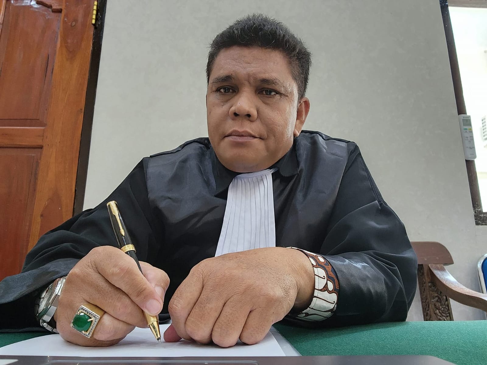 Potret Raymond Simamora, S.H., M.H. yang tampak fokus saat bersiap menandatangani dokumen hukum dengan mengenakan atribut toga advokat lengkap di meja persidangan.